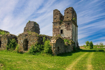 Crumbling stone towers and walls of the historic Pidhoriansky Monastery near Terebovlia, Ukraine. Overgrown ruins set against a vibrant blue sky with wispy clouds and lush green meadow