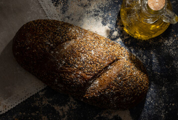 Top view of artisan whole grain rye bread next to a glass jar of oil on a dark wooden background...