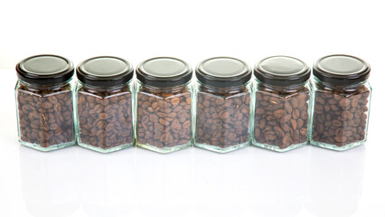 six glass jars with black lids filled with roasted coffee beans arranged in a row on a reflective surface on a white background