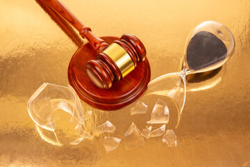 Close-up of a polished gavel next to a broken hourglass with dark sand, on a golden surface,...