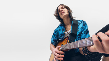 Young man, guitarist in tropical shirt strumming melody with closed eyes, deeply connected to music, playing guitar on white background. Concept of music, festival, performance, concert, rock, talent