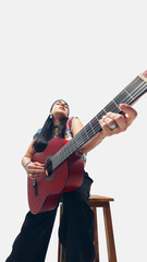 Fototapeta premium Hippie-style guitarist, woman in colorful vest and headband passionately playing red guitar, deeply engaged in sound. Low angle view. Concept of music, freedom, folk, art, cultural expression