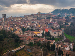 Fototapeta premium The old town of Bergamo, Italy, at sunset features terracotta roofed buildings, a bell tower, a dome, lush greenery, and rolling hills under dramatic clouds.