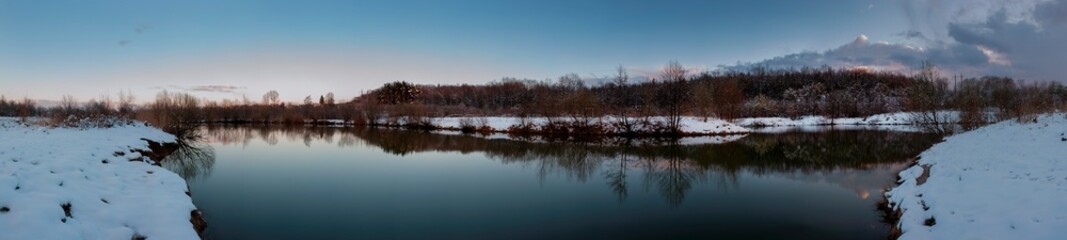 Fototapeta premium Panorama of the sunrise over the winter river. The sun rises over the lake. Autumn frost. The frozen river is covered with thin ice, the grass glistens with frost.
