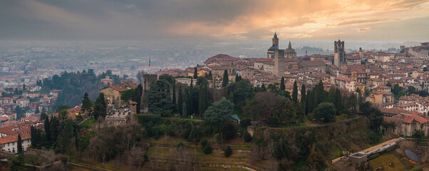 View of Bergamo's Citta Alta, featuring the Campanone, Basilica of Santa Maria Maggiore, terracotta rooftops, and Italian flag at sunset.