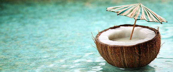 Half coconut shell with coconut water, topped with a small striped umbrella, floats on a turquoise surface, showcasing a refreshing summer drink concept