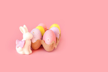 Eco basket with colorful easter eggs with white rabbit or bunny statuette. Minimal pastel light pink background. minimalistic decoration, composition. Front view, place for text, banner