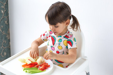 Little girl child 2-3 year with pigtails sitting in high chair, picking fresh vegetables from plate. Healthy eating habits, childhood nutrition, and natural food choices