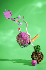 Pink watering can pouring flowers onto a moss ball balanced by a gardening tool on a decorative branch, set against a vibrant green background, embodying spring and growth