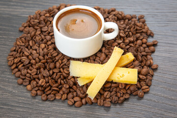 A top-down view of a white cup of rich espresso on a saucer, accompanied by yellow cheese sticks, surrounded by roasted coffee beans on a dark wooden surface