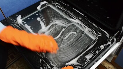 Person is cleaning a stove top with a sponge and soap. The stove top is covered in a white substance that looks like soap suds. The person is wearing an orange glove while cleaning