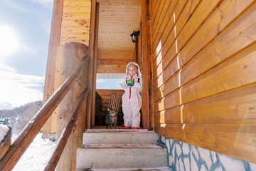 Little girl stands with a tabby cat on the threshold of a wooden cottage in a snowy village and...