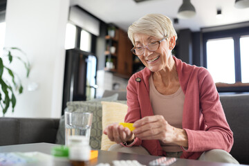 Smiling senior woman sitting at home holding a pill bottle and preparing to take medication with a...