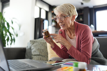 Senior woman sitting at home holding a pill bottle while consulting a doctor via video call on her laptop. Concept of telemedicine, online healthcare, and medication management for seniors