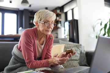 Senior woman sitting at home holding a pill bottle while consulting a doctor via video call on her laptop. Concept of telemedicine, online healthcare, and medication management for seniors