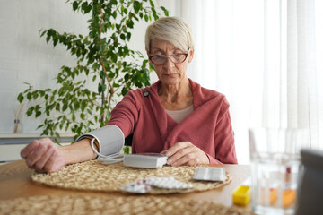 Senior woman measuring her blood pressure at home using a digital monitor. She looks focused while...