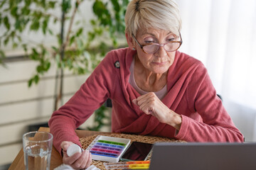 Senior woman using a laptop while holding a pill bottle, engaging in a telemedicine consultation or researching medication online. Concept of digital healthcare and senior medical awareness.