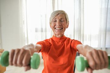 An elderly woman sits on a chair, lifting green dumbbells while smiling. She exercises in a bright...