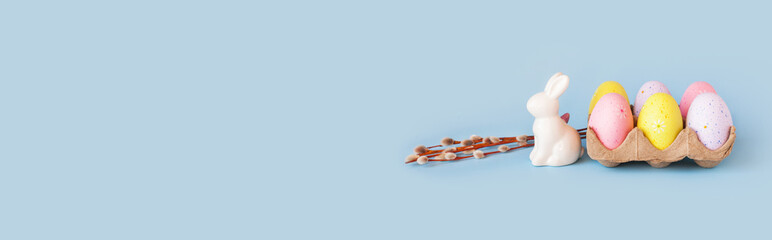 Happy easter decoration. White bunny decorative statuette, willow branch, basket with colorful eggs. Minimal festive composition on pastel light blue background. Copy space, place for text, front view