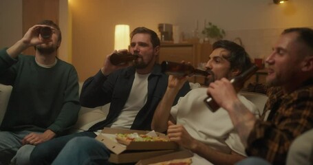 Four happy friends sitting on the sofa clinking beer bottles and eating pizza during a bachelor party at home in the evening