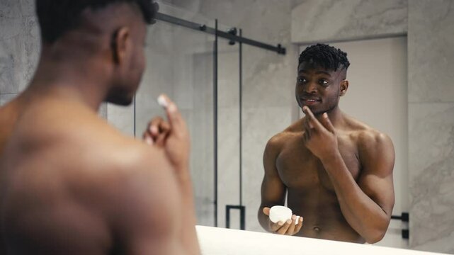 Young African American man applying cream in front of a mirror, skincare routine