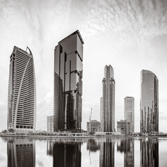 Fototapeta premium Monochrome city skyline with tall skyscrapers reflecting on calm water under a cloudy sky, Dubai Marina bay UAE