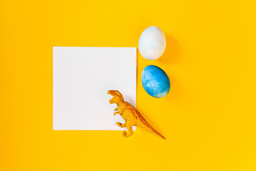 Easter egg hunt holiday composition, paper note or list with colorful eggs, dinosaur on yellow backgroind. Seasonal spring kids play concept. Flat lay, top view, place for text