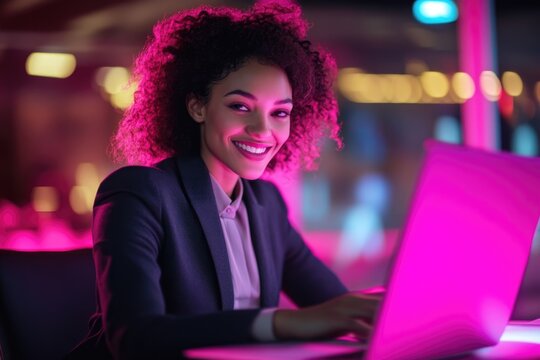 A smiling businesswoman works on her laptop in a vibrant, brightly lit office at night.