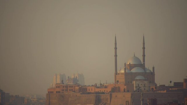 The Mosque of Muhammad Ali stands atop Cairo Citadel, its domes and towering minarets illuminated by soft haze. Modern high-rises loom in the distance, contrasting the historic fortress below.