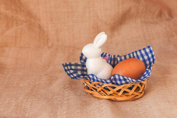 Happy easter decoration with white bunny decorative statuette, egg and willow branch in basket. Minimal isolated festive composition on eco textile background. Copy space, place for text, front view