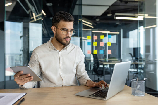 Multitasking, man with tablet computer working inside office using laptop. Businessman serious and focused typing on keyboard. Company employee thinking and concentrated. - Powered by Adobe