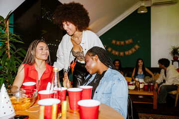 A woman stands next to two friends sitting at a table, they are laughing, in the living room, at a party