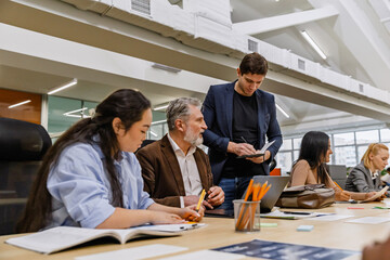 A male employee stands and writes in a notebook while a group of four employees sit at a table next...