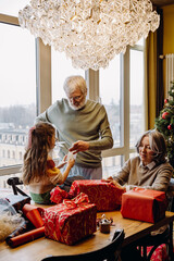 Family, grandfather standing and cutting tape which he holds together with granddaughter sitting next to him on the table while grandmother sits next to them, in the living room with Christmas tree