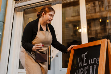 A White woman in her late 20s, a restaurant worker, in a beige apron and a black shirt, is writing on a blackboard, standing in front of the entrance doors of the restaurant, outside, during daytime.