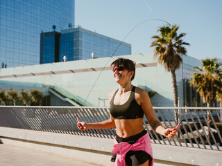 A smiling adult White woman in sunglasses and a black top jumps on a jumping rope, smiling on a sunny day. The woman exercises on a bridge with metal railings with modern buildings in the background.