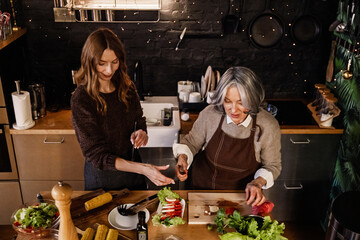 A gray-haired European mother in her early 50s in a brown apron cooking with her dark-haired daughter in her early 30s in a brown sweater reaches out to her, standing in the kitchen.
