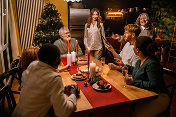 A group of multiethnic adult people of various ages sits and stands at a wooden table with food, champagne flutes, and candles in a kitchen with a Christmas tree, preparing to eat and celebrate.