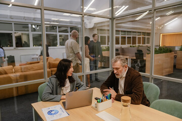 An Asian woman in her 30s and a White middle-aged man sitting at a table in the office with glass walls and talking, a multiethnic group of colleagues standing in the background in the office hall