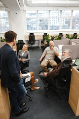 A formally dressed brunet White man in his 30s stands and talks to his formally dressed White colleagues who sit at their desks, and look at him, in a light office with windows in the background.