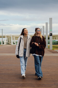 Two young White women in their 20s walking on a wooden pathway outdoors, wearing casual winter outfits. One wears a white fleece jacket, while the other wears a brown sweater and sunglasses.