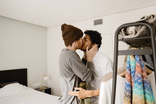 A multiracial gay couple shares a kiss in a cozy bedroom. One man wears a beanie and casual clothes, while the other wears a white shirt. The intimate setting features a bed and a clothing rack.
