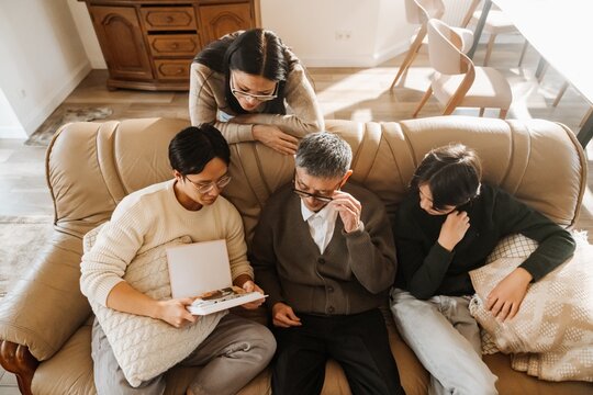 An Asian multigenerational family sits on a leather couch, looking at a photo album. The elderly grandfather adjusts his glasses while his children and grandchildren engage with the memories.