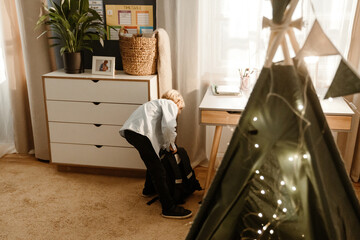 Fototapeta premium A white-skinned boy in a light blue shirt and black pants bends down to pack his backpack in a cozy bedroom. A study desk with books and pencils, a dresser, a timetable are visible.