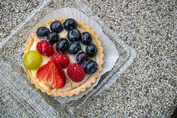 A sweet tart in a plastic box lies on a stone surface, Shortcrust pastry with cream, half a strawberry, blueberries and raspberries lie on top of the cream, covered with clear syrup.