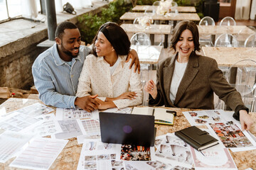 An adult Black man and a woman sit together at a table with a White female wedding consultant. The table is covered with bridal posters and a laptop. The concept of love, marriage, and organization.