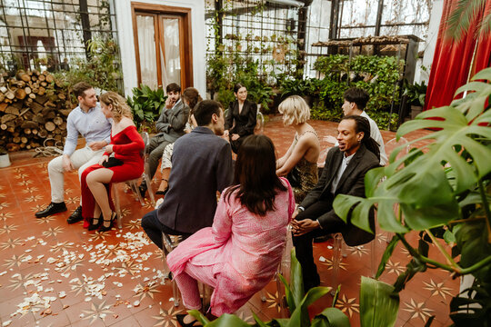 A multiethnic group of elegantly dressed guests sits in a circle, engaged in conversation at a wedding reception. The venue features lush greenery, wooden decor, rose petals scattered on the floor. - Powered by Adobe