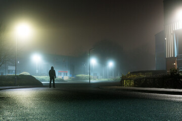 A mysterious sinister figure. Standing on a road, on an industrial estate with warehouses. On a spooky foggy winters night.