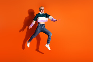 Cheerful young woman in casual clothing jumping joyfully against vibrant orange background, spreading positivity