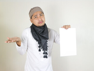 handsome asian muslim man wearing black turban or shemagh holding blank white paper and showing...